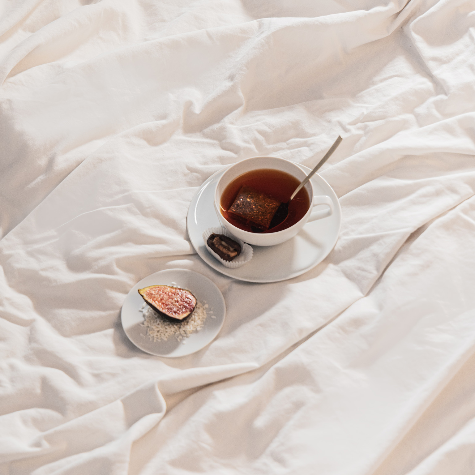 La tasse à thé TAC remplie de thé, la soucoupe et l'assiette à pain avec une figue sont posées sur un drap de lit.